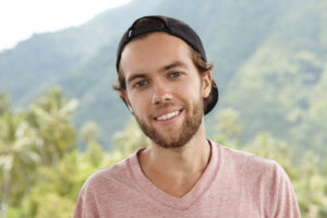 Outdoor shot of young Caucasian man with beard relaxing in open air, surrounded by beautiful mountain setting and rainforest, looking at camera and smiling happily during vacations in exotic country